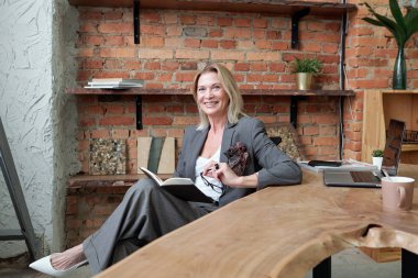 Gülümseyen, sarı saçlı, gri takım elbiseli bir iş kadını ofiste oturmuş kitap okuyor.