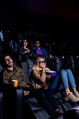 Üç boyutlu gözlüklü kız arkadaşı patlamış mısır yiyip sinemadan zevk alırken, yorgun adam sinemada koltukta uyuyor.