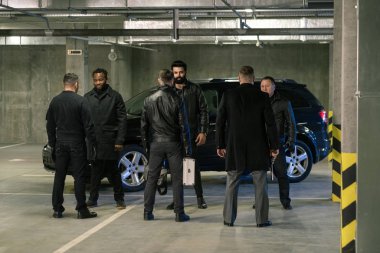 Siyahlı birkaç kültürler arası adam park alanında suç işleme şartlarını tartışırken tabanca ve bavul taşıyorlar.