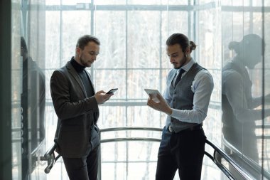 İki iyi giyimli iş adamı ara verdiğimizde modern iş merkezinin penceresinde beklerken cep aletlerini kullanıyorlar.