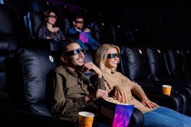 Üç boyutlu gözlüklü genç çift büyük ekranın önünde oturmuş film izlerken sinemada eğleniyor.