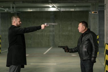 Siyah ceketli ve pantolonlu iki yetişkin birbirlerinin önünde tabancayla ateş etmeye hazırlar.