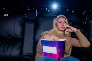 Sıkıcı sarışın kız elinde patlamış mısırla sinemada büyük ekranın önünde siyah deri koltukta oturup film izliyor.