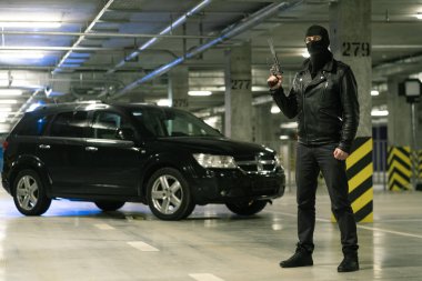 Siyah ceketli bir terörist ya da gangster ve kafasında kar maskesi olan tabanca, arka planda arabayla vurmaya hazır.