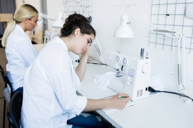 Kafası karışmış beyaz önlüklü genç bir kadın eskiz defterindeki notlara bakarken fabrika atölyesindeki dikiş kalıplarını düşünüyor.