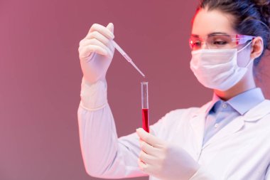 Laboratuvarda deney yaparken koruyucu gözlüklü ve maskeli genç bir kimyager kırmızı sıvıyı şişeye boşaltıyor.