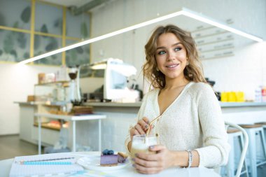 Cafe 'de masa başında oturmuş, kapuçino ve lezzetli yaban mersinli kek yiyen hoş, güler yüzlü bir kadın.