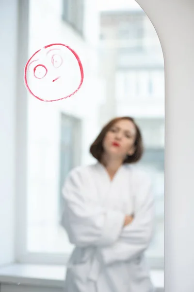真紅の口紅で描かれた空白の顔を持つ鏡の前に立つ白いバスローブの若い武装横断型の女性 — ストック写真
