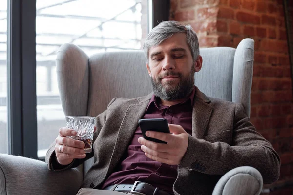 Content gray-haired businessman with beard sitting with whisky glass and scrolling in smartphone