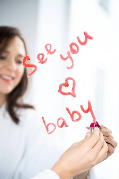 Hand of brunette woman writing love message and drawing heart with red lipstick on mirror after morning hygiene