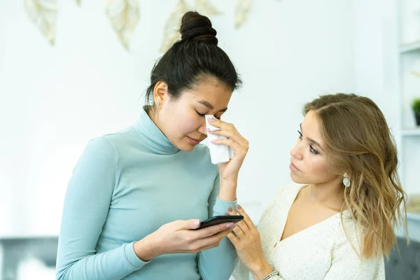 Pretty young woman comforting her upset friend crying after getting bad message on smartphone