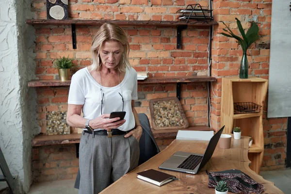 Serious attractive lady boss checking phone message while standing in modern office