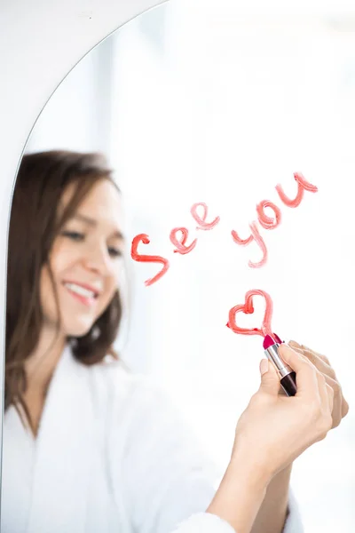 Hand of happy young female in white bathrobe writing see you and drawing heart with lipstick on mirror