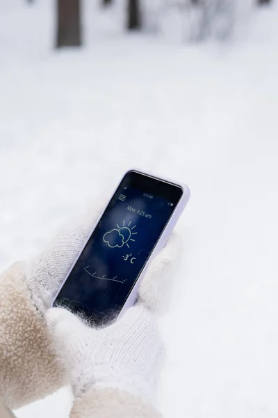 Hands of girl in white fluffy knitted mittens looking for temperature outside in mobile phone while chilling in snowbound park