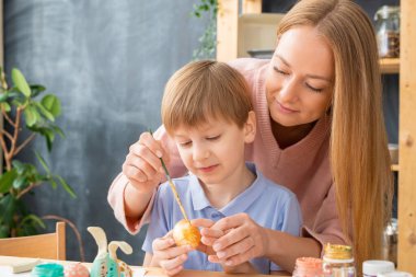 Paskalya süslemeleri için yumurta boyamasına yardım ederken boya fırçası kullanan çekici genç bir anne.