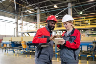 Content young multi-ethnic coworkers in hardhats using tablet while discussing manufacturing issues at construction plant