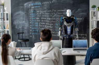 Otomasyon robotunun önünde oturan öğrencilerin arka görünümü insan öğretmen yerine karatahtanın yanında duruyor.