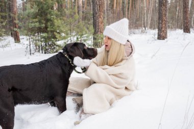 Uzun sarı saçlı, karda oturmuş labrador köpeğiyle eğlenen genç kadının yatay görüntüsü.