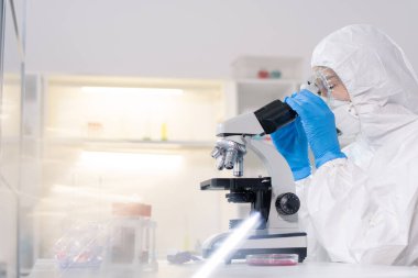 Focused lab technician in protective suit and mask using microscope while working with blood samples