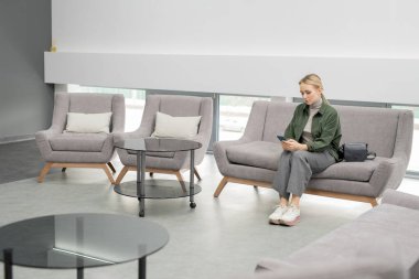 Günümüz tıp merkezindeki koltukta oturan ve sırasını beklerken telefonu kullanan gündelik kıyafetli genç sarışın kadın.