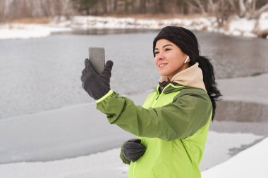 Smiling pretty woman in ear buds using smartpnone while taking selfie against winter lake