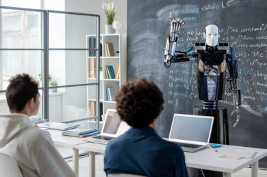 Otomasyon bilgisayar robotuna bakan iki erkek öğrenci ders sırasında teknik verilerle karatahtanın yanında duruyor.