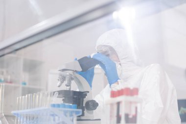 Behind glass view of virology scientist in protective suit analyzing blood sample under microscope