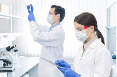 Two professional pharmacologists wearing protective masks, gloves and eyeglasses working on medicaments in modern laboratory