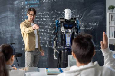 Ciddi bir öğretmen, öğrencilerden birinin yanında duran otomasyon robotunun teknik özelliklerini saymasını istiyor.