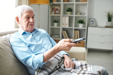 Kareli ekoseli yaşlı adam evde televizyon izlerken koltukta oturup uzaktan kumandaya basıyor.