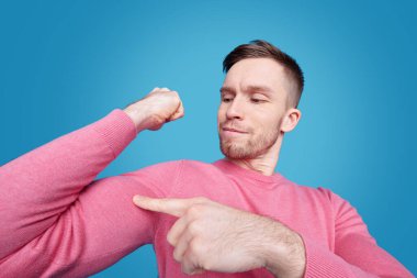 Pembe kazaklı, mavi arka planda kaslarını esneten kendinden emin genç sakallı bir adam.