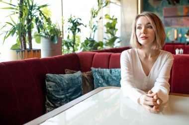 Sarışın kadın modern kafede tek başına oturmuş birini beklerken bir şeyler düşünüyor, yatay portre.