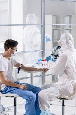 Doctor in protective suit taking blood of Asian coronavirus patient while undergoing medical research in laboratory
