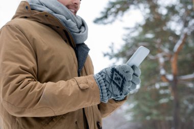 Genç bir adam kış günü açık havada dikilirken akıllı telefonuyla, yatay çekim yapıyor.