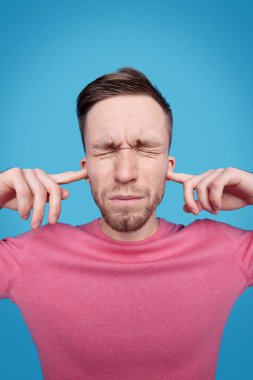 Sinirli, sakallı, gözleri kapalı genç bir adam kulakları mavi arka plandaki yüksek seslerden koruyor.