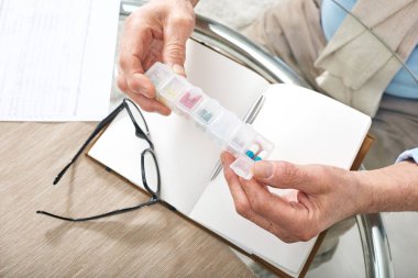 İlaç almaya giderken masanın üzerinde plastik hap kutusu ve gözlük taşıyan erkek emeklilerin elleri.