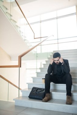 Kafası karışmış, siyah takım elbiseli genç bir iş adamı elinde kahve fincanıyla merdivenlerde oturuyor ve elini tutuyor.