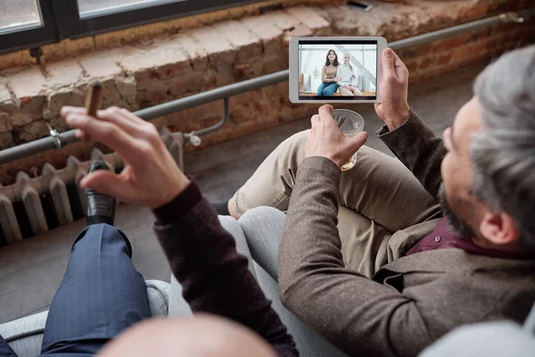 Over shoulder view of businessman with glass sharing photo of wife and daughter on tablet to friend at party