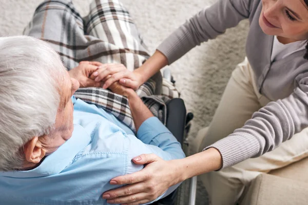 Young supportive female caregiver sitting by senior man in wheelchair and keeping her hand on his shoulder while comforting him