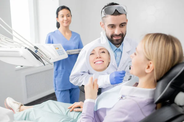 歯医者のオフィスで椅子に座っている間にプロのホワイトニングの手順の後に鏡を見て若い笑顔の女性 — ストック写真