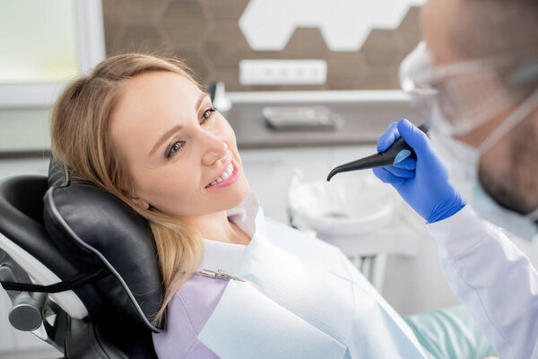 Красивая молодая блондинка с головой на черной кожаной подушке глядя на дантиста собирается починить зубы
