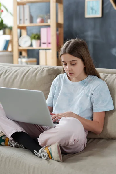 严肃的少女穿着家居服 双腿交叉坐在沙发上 上网聊天时使用笔记本电脑 — 图库照片