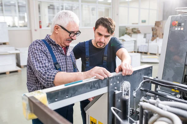 在现代工厂 戴眼镜向年轻同事解释印刷机装置的资深工人 — 图库照片