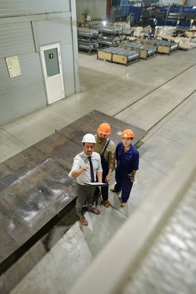 同僚にそれを示しながら 新しい巨大な産業機械を指している制服とハードハットの3人の若いエンジニアの一人 — ストック写真