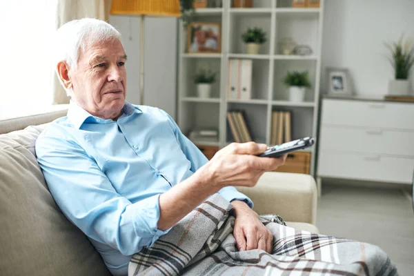 老人坐在沙发上 边看电视边按遥控器上的按钮 — 图库照片