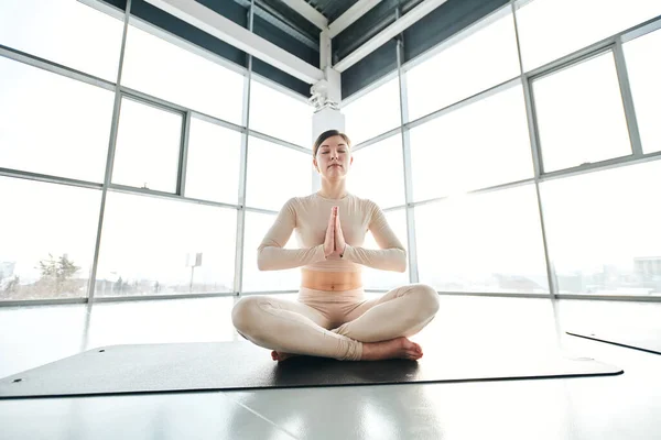 Serene girl in activewear sitting in pose of lotus on mat during relaxation exercise on background of large window in gym