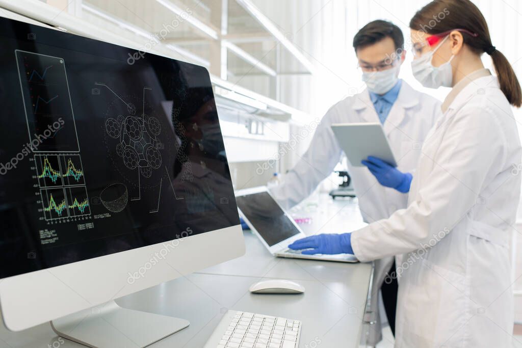 Horizontal shot of two scientists wearing white coats, masks and gloves working in laboratory using computers and gadgets