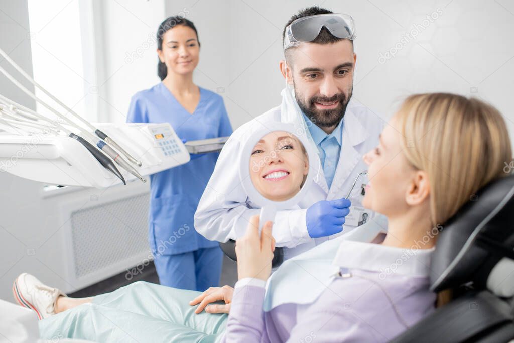 Young smiling woman looking in mirror after procedure of professional whitening while sitting in armchair in the dentist office