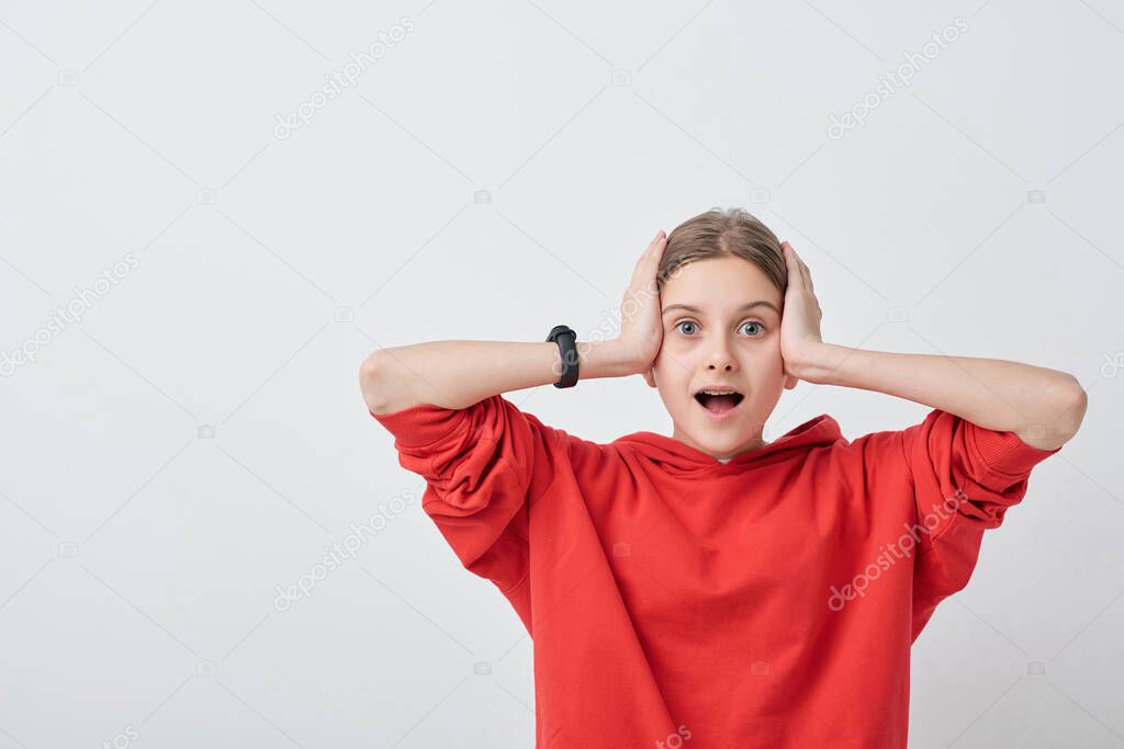 Portrait of shocked teenage girl in red hoodie keeping mouth open and holding head in hands against white background