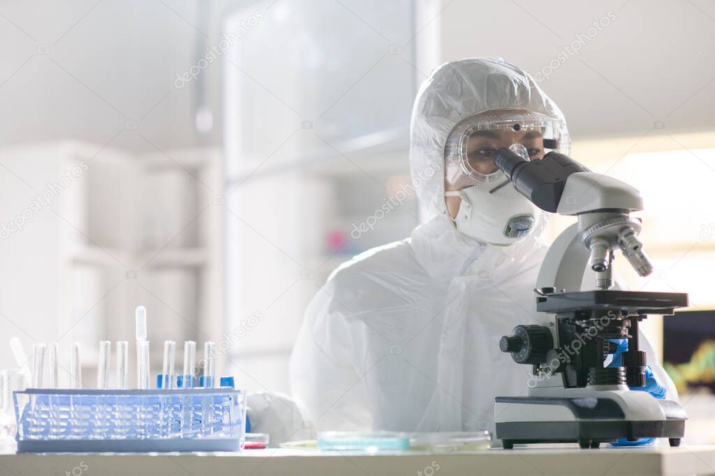 Concentrated Asian virology researcher in protective suit using microscope while testing virus sample in laboratory